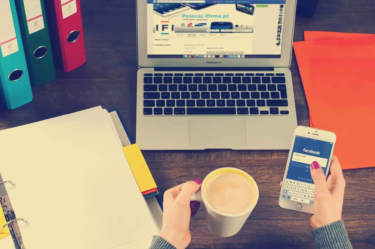 Social media manager working with laptop and phone, managing Facebook and other social platforms from a single workspace